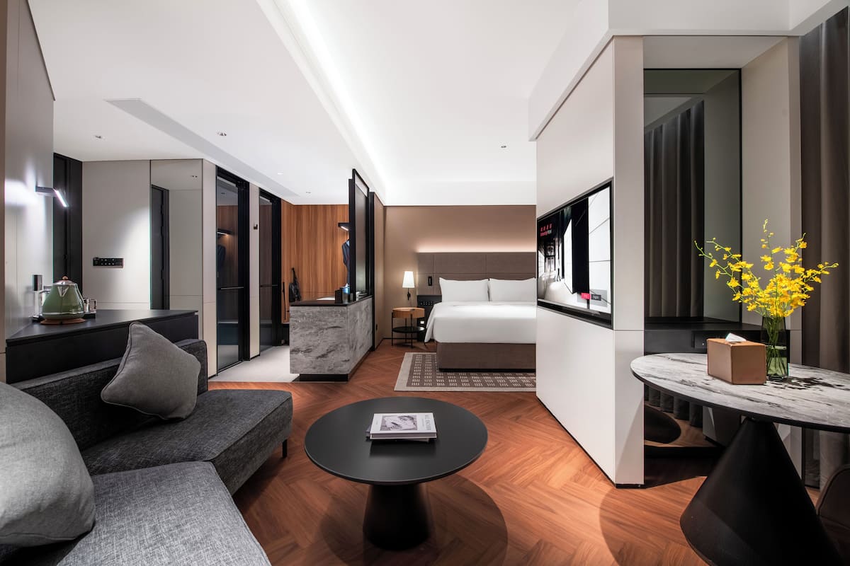 Una habitación de hotel moderna con un sofá, una mesita redonda, una cama, un televisor y un jarrón con flores amarillas.