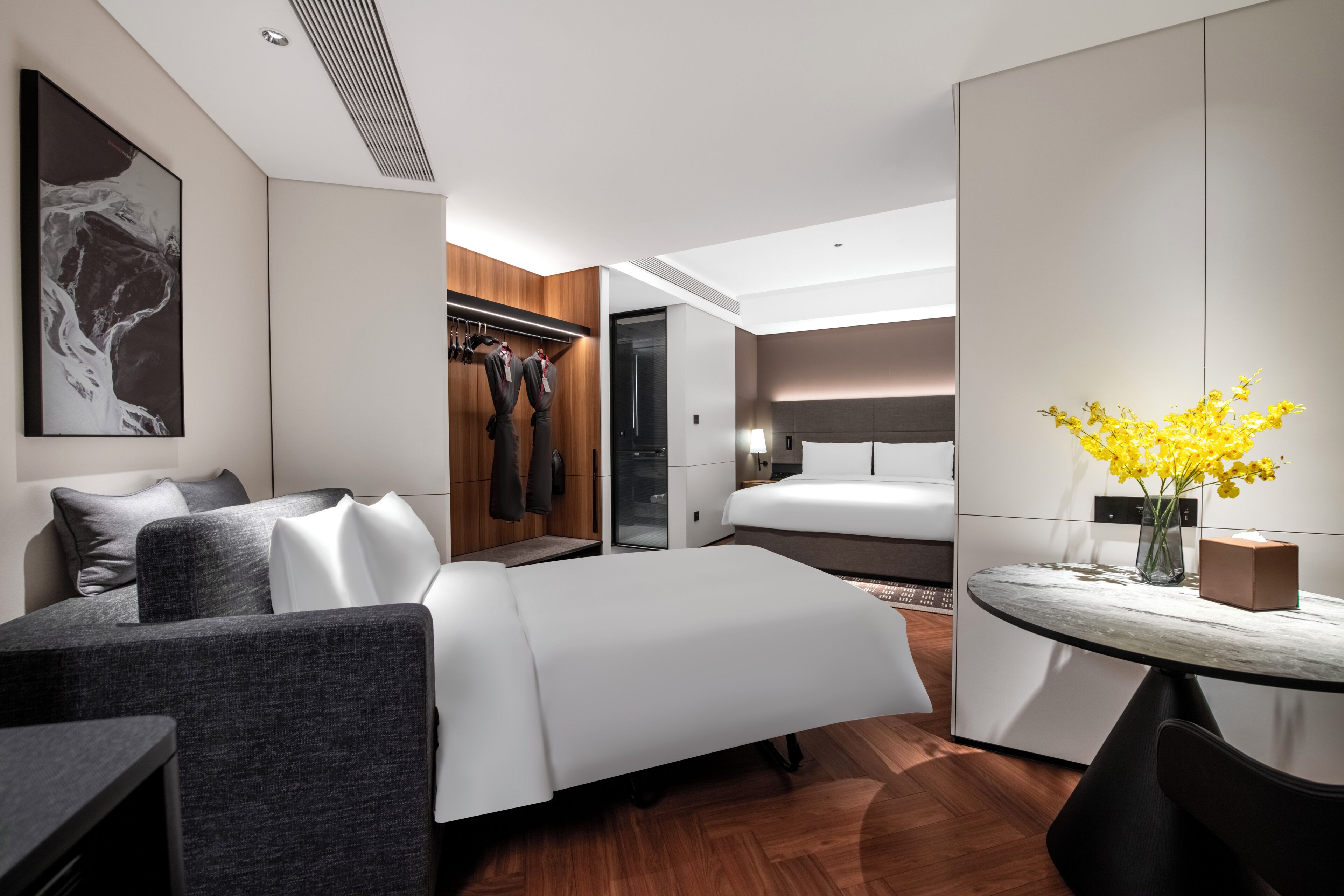 A modern hotel room with a bed, seating area, wardrobe, and a dining table with flowers.