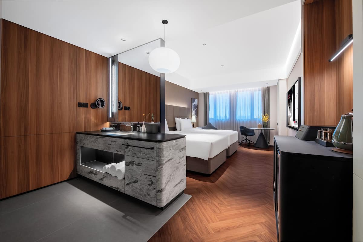 Habitación de hotel moderna con tocador de mármol, cama con ropa blanca, escritorio con silla y un amplio ventanal con cortinas.