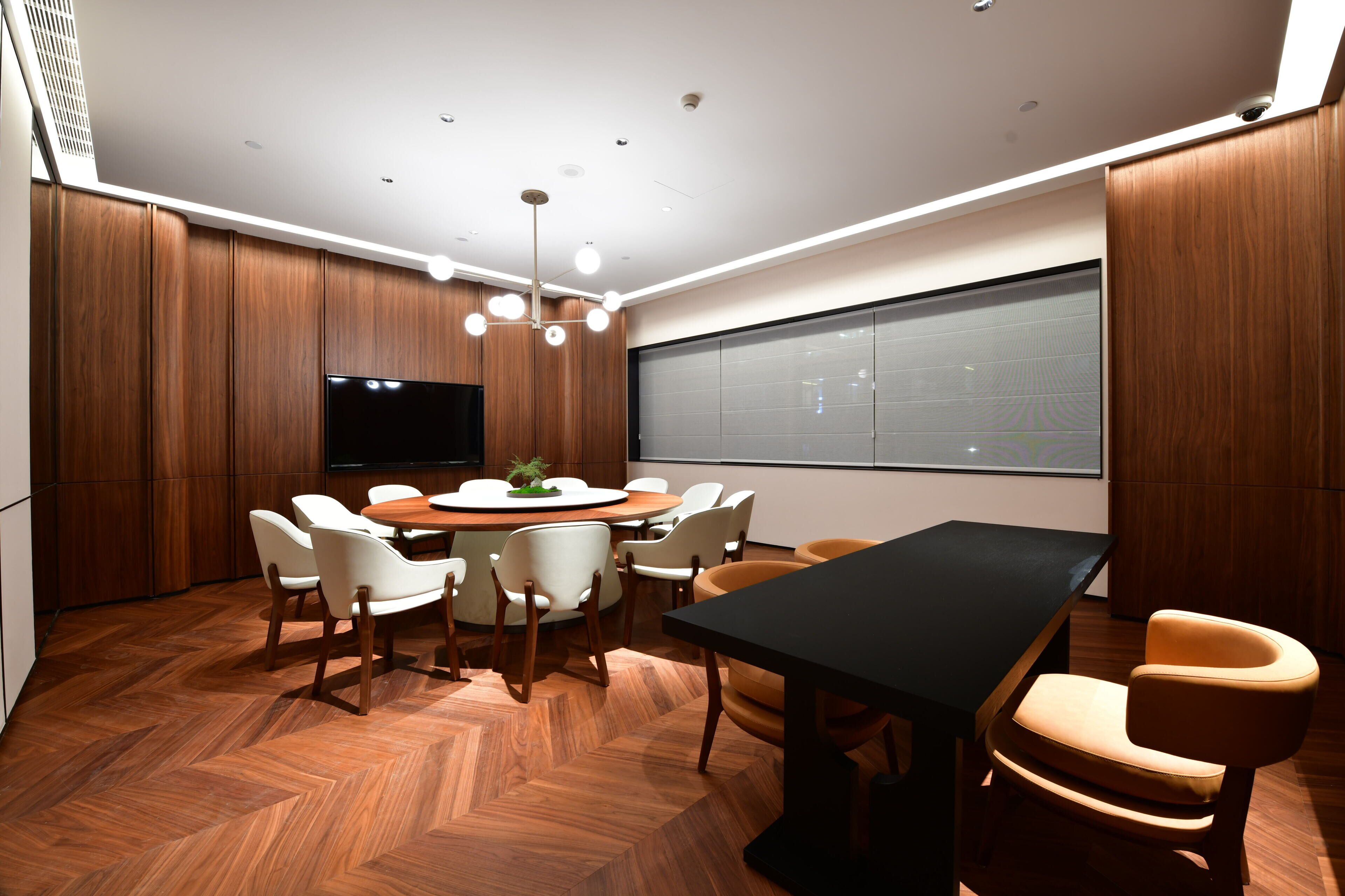 A conference room with a round table, white chairs, a black table, and a large screen.