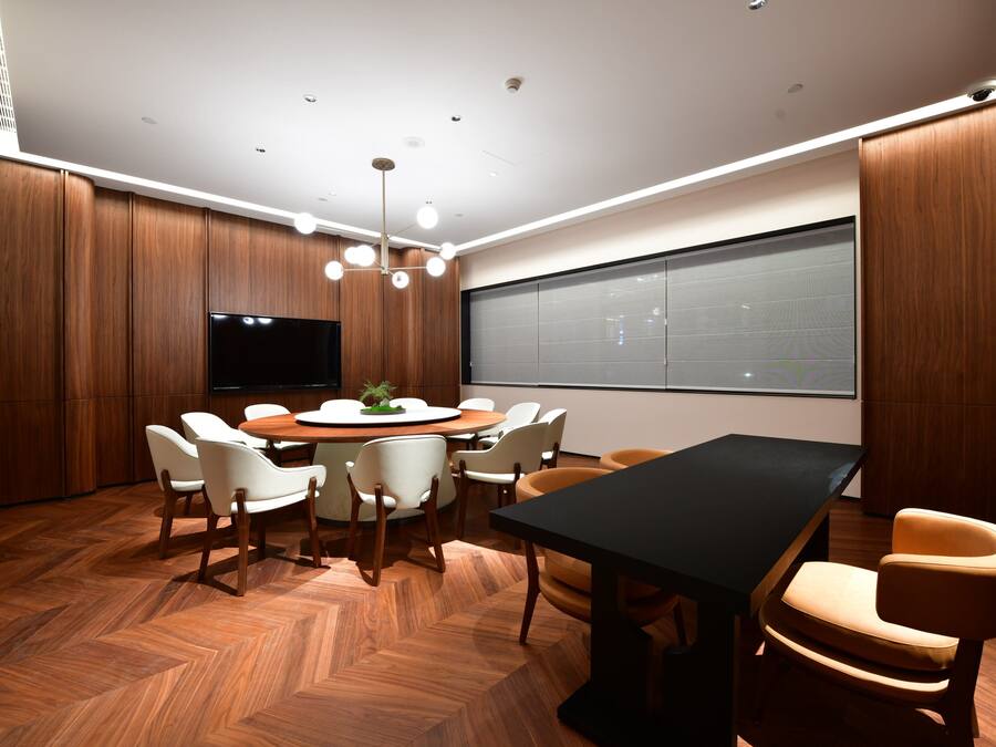 A conference room with a round table, white chairs, a black table, and a large screen.