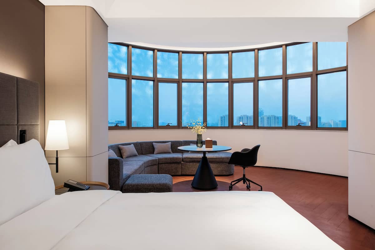 Una habitación de hotel moderna con un ventanal curvo, un sofá, una mesa redonda y una cama.