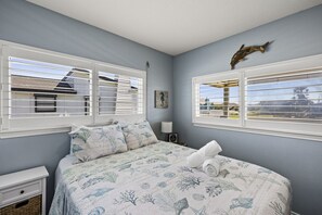 Une chambre à coucher avec un lit, deux fenêtres, une table de chevet, une lampe et un élément décoratif fixé au mur.