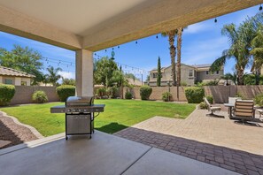 A covered patio with a grill, outdoor furniture, and a view of a well-maintained lawn and neighboring houses. - Family-Friendly 4BR Home, 30+ Night Stays -  Loft, Big Backyard (Gilbert)