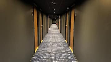 Un couloir d’hôtel avec un sol à carreaux, des portes de chaque côté et un extincteur accroché au mur.