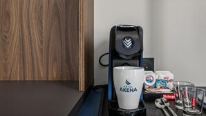 A coffee machine, a white mug with 'HOTEL AKENA' logo, a spoon, a glass, and a box of Malongo coffee pods on a table.