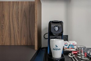 Eine Kaffeemaschine, eine weiße Tasse mit dem Schriftzug 'HOTEL AKENA', ein Löffel, ein Glas und eine Schachtel Malongo-Kaffeekapseln auf einem Tisch.