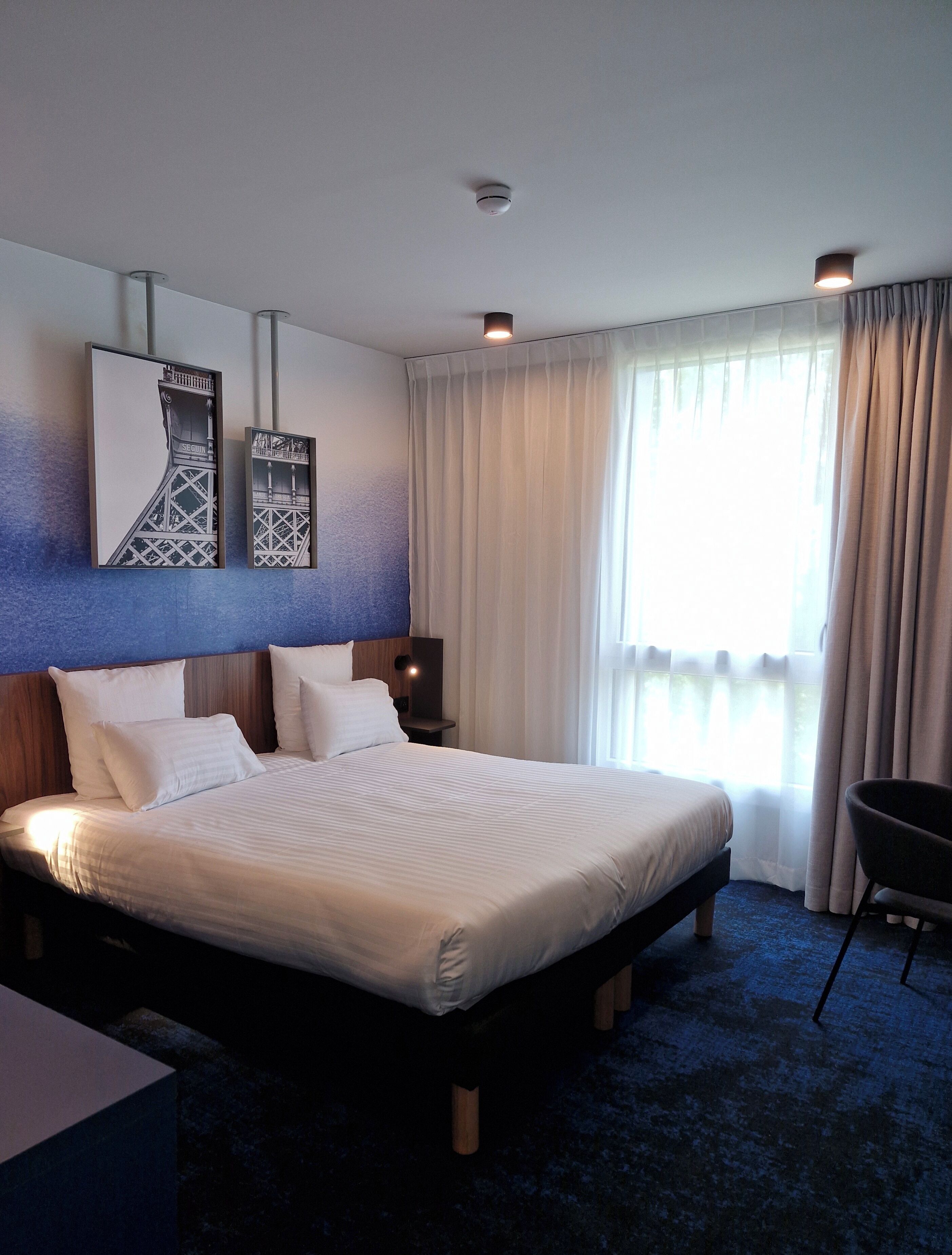 A hotel room with a large bed, a headboard, a nightstand, a chair, and two wall-mounted artworks.