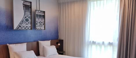 Une chambre d’hôtel avec un grand lit, une tête de lit ornée d’une œuvre d’art, une table de chevet avec une lampe, une chaise et une fenêtre avec des rideaux.