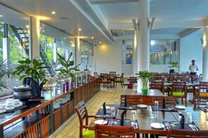 A spacious dining area with multiple tables and chairs, a buffet counter with food and drink dispensers, and large windows offering views of greenery outside. - Raintree by Oak Ray - Tissamaharama (Ella)