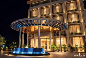 A hotel entrance with a covered outdoor area, a water feature, and a building with multiple floors and balconies.
