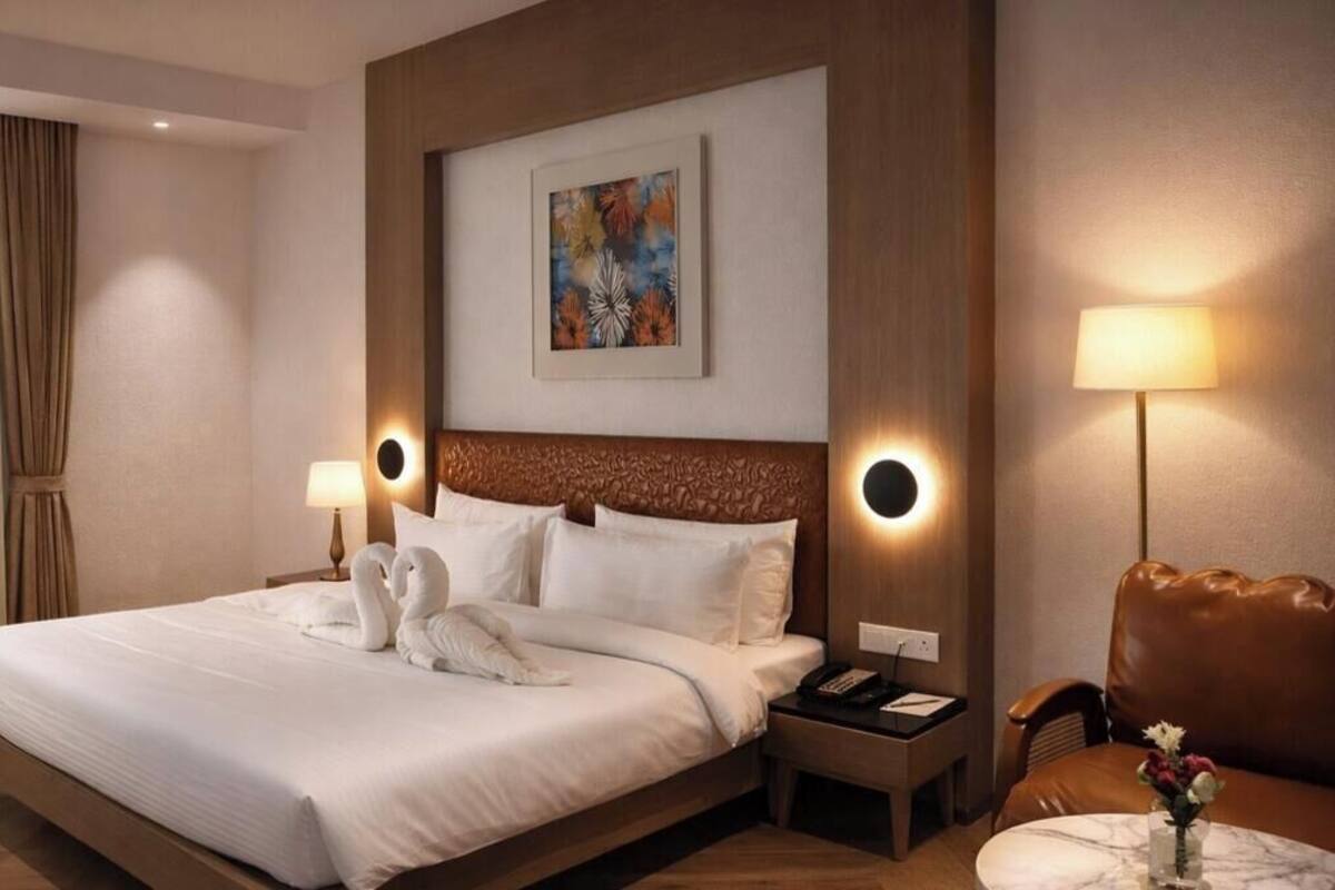 A hotel room with a large bed, two bedside lamps, a headboard with a circular light, a painting on the wall, and a small table with a vase of flowers.