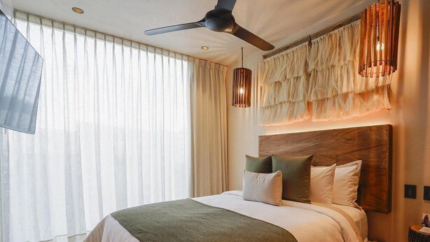 A hotel room with a large bed, a ceiling fan, a wooden headboard, and a large window with curtains. - Trobbu (Tulum)