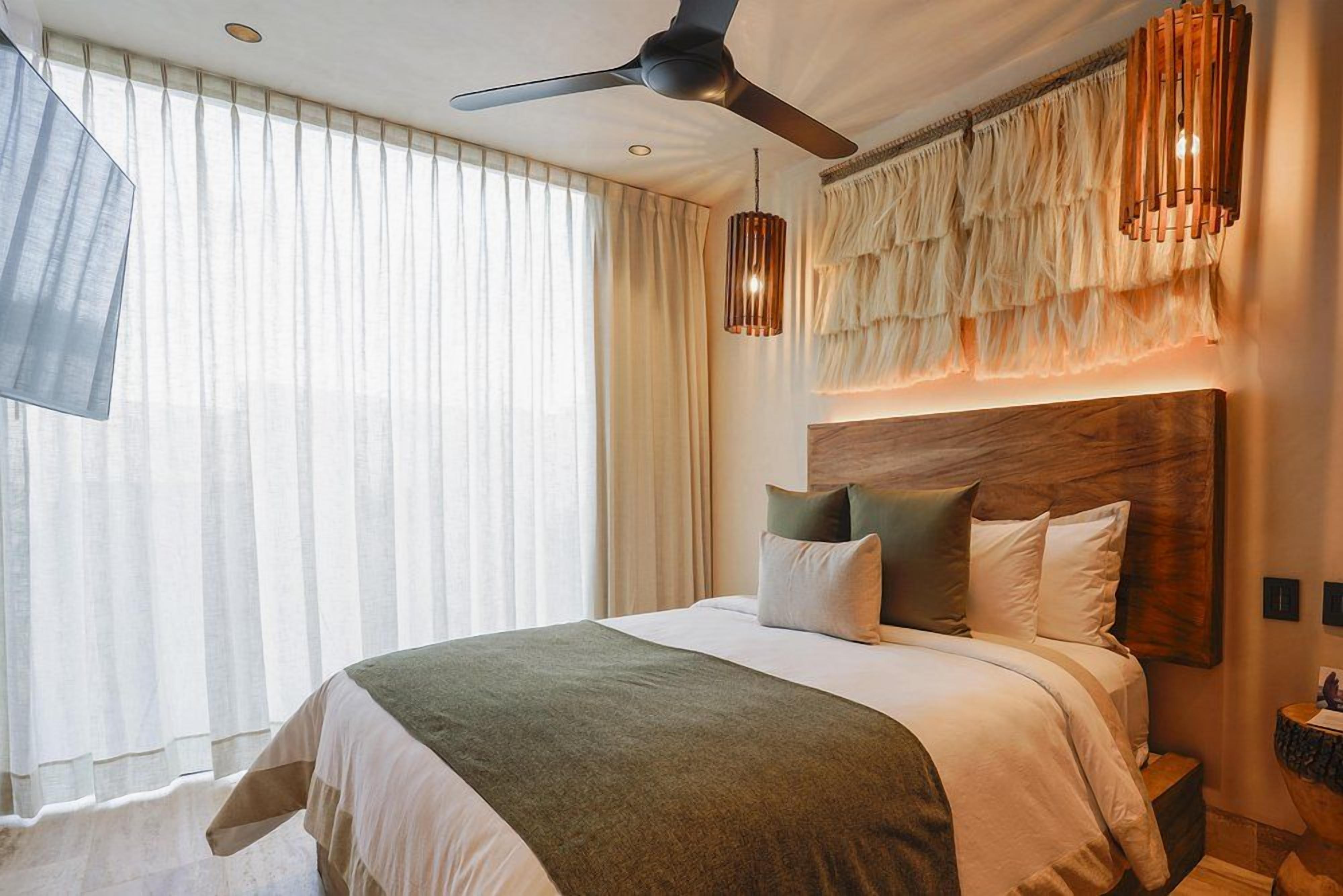 Habitación de hotel con una cama grande, ventilador de techo, cabecero de madera y un amplio ventanal con cortinas.