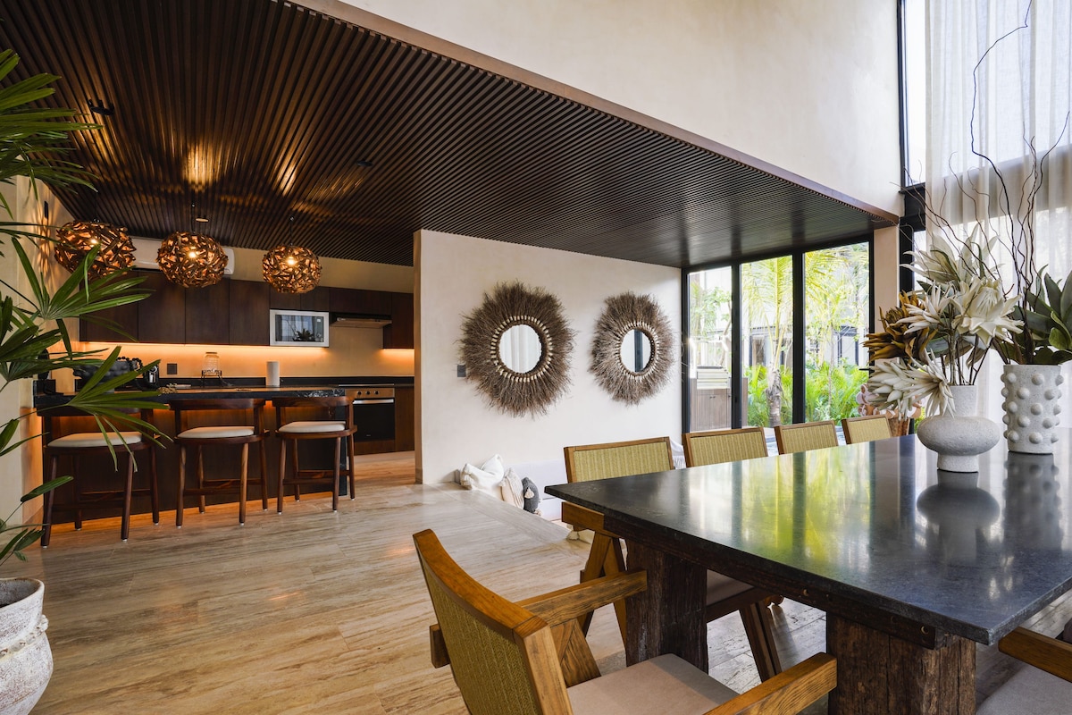A modern dining area with a large table, wooden chairs, and a kitchen in the background.