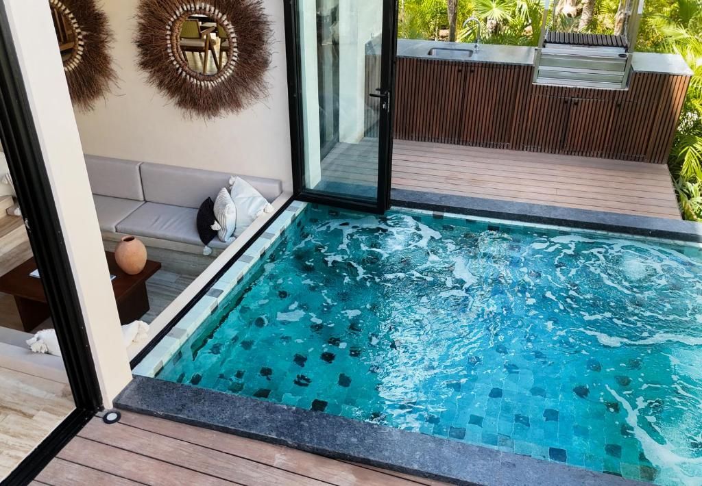 Una piscina con borde de azulejos, terraza de madera, puerta de cristal que da a un balcón y un sofá con cojines.