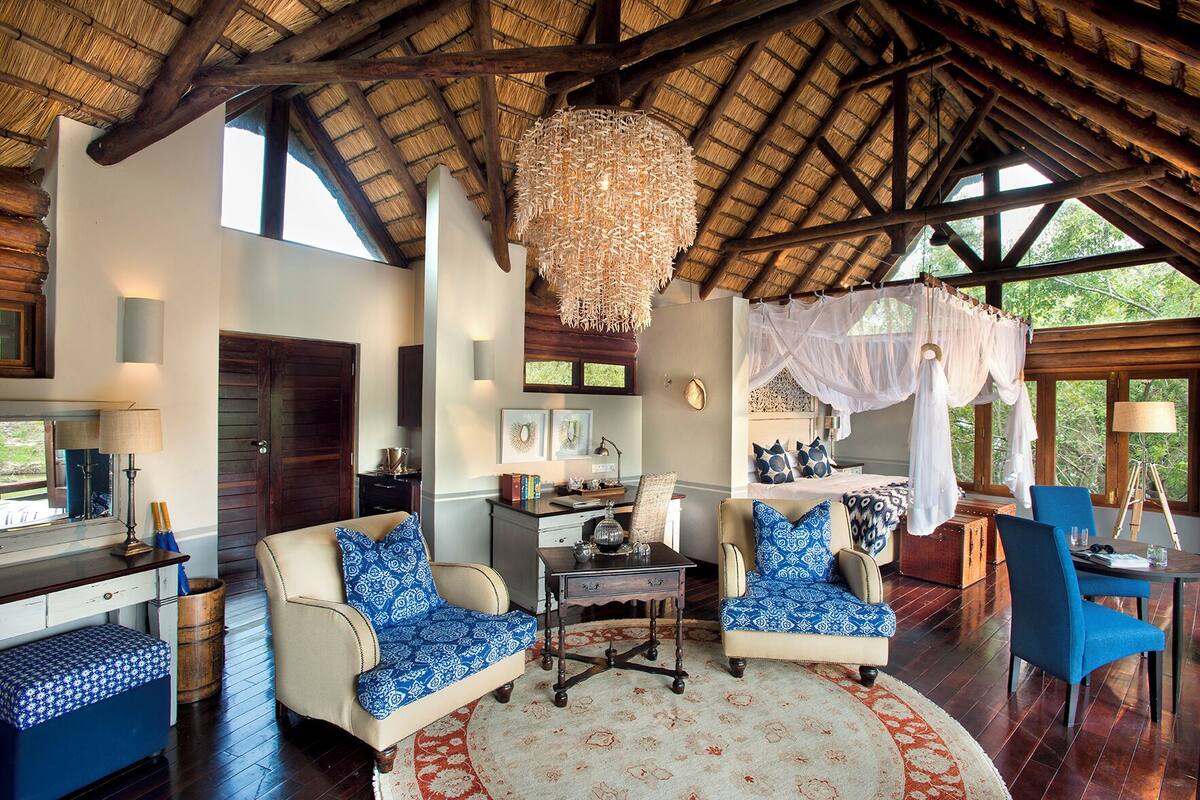 A spacious room with a high ceiling, a large chandelier, a bed with a canopy, and a dining area with blue chairs.