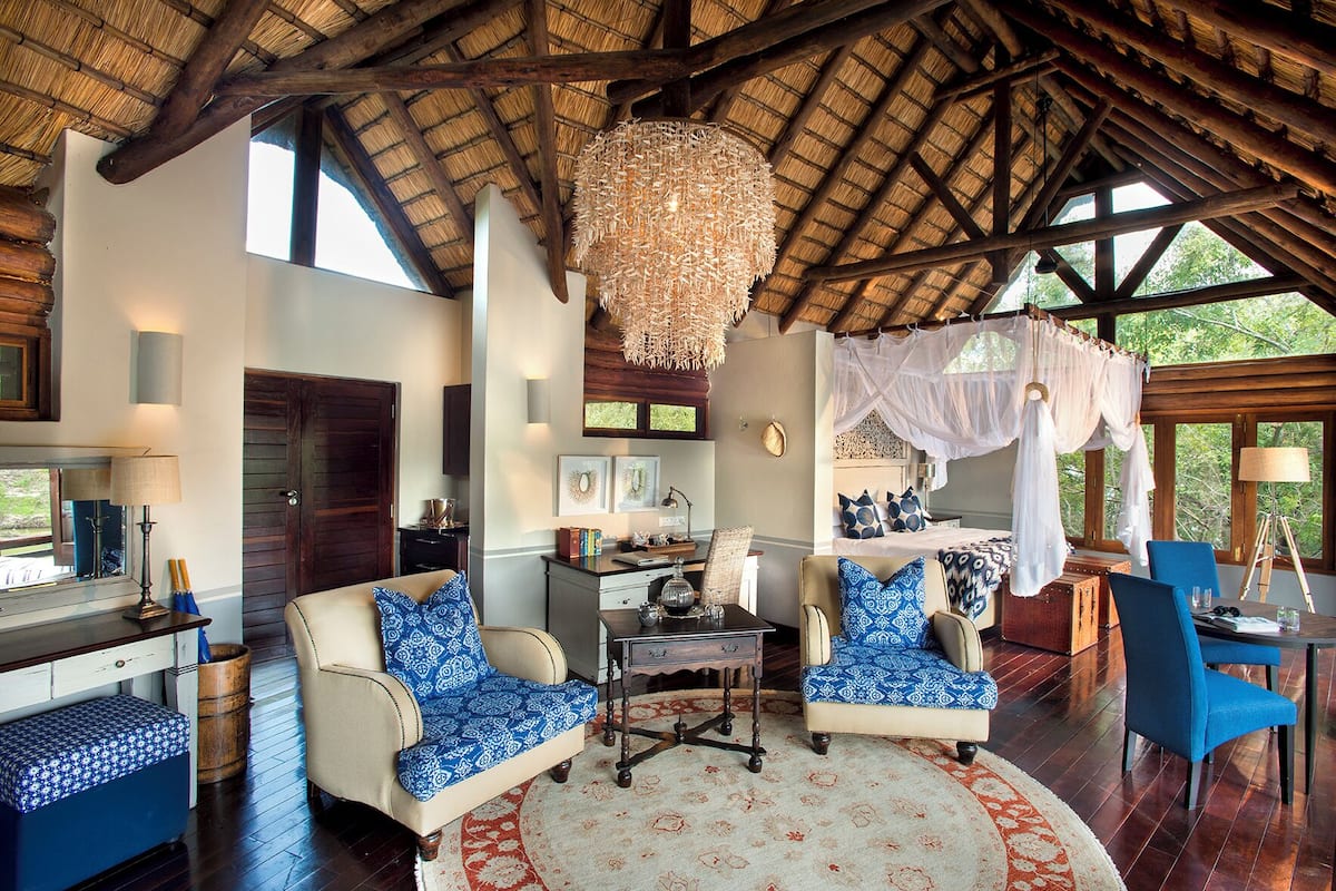 A spacious room with a high ceiling, a large chandelier, a bed with a canopy, and a dining area with blue chairs.