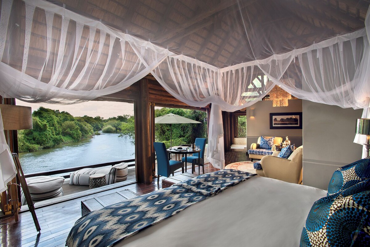 A bedroom with a large bed, a sitting area with a table and chairs, and a view of a river.
