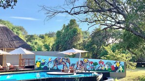 Une piscine ornée d’une fresque murale, dotée de chaises longues et surmontée d’une structure à toit de chaume.