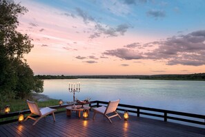 Una terraza con dos sillas, una mesa y un candelabro con velas, con vista a un cuerpo de agua tranquilo y un cielo al atardecer.