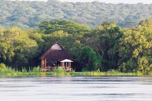 A thatched-roof hut with a canopy, surrounded by lush greenery and a calm body of water. - Royal Chundu River Zambezi Lodge (Livingstone)
