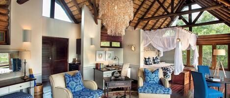 A spacious room with a high ceiling, a large chandelier, a bed with a canopy, and a dining area with blue chairs.
