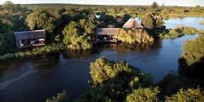 Un chalet au bord de la rivière, doté de bâtiments à toits de chaume, entouré d’une végétation luxuriante et bordé par un fleuve paisible.