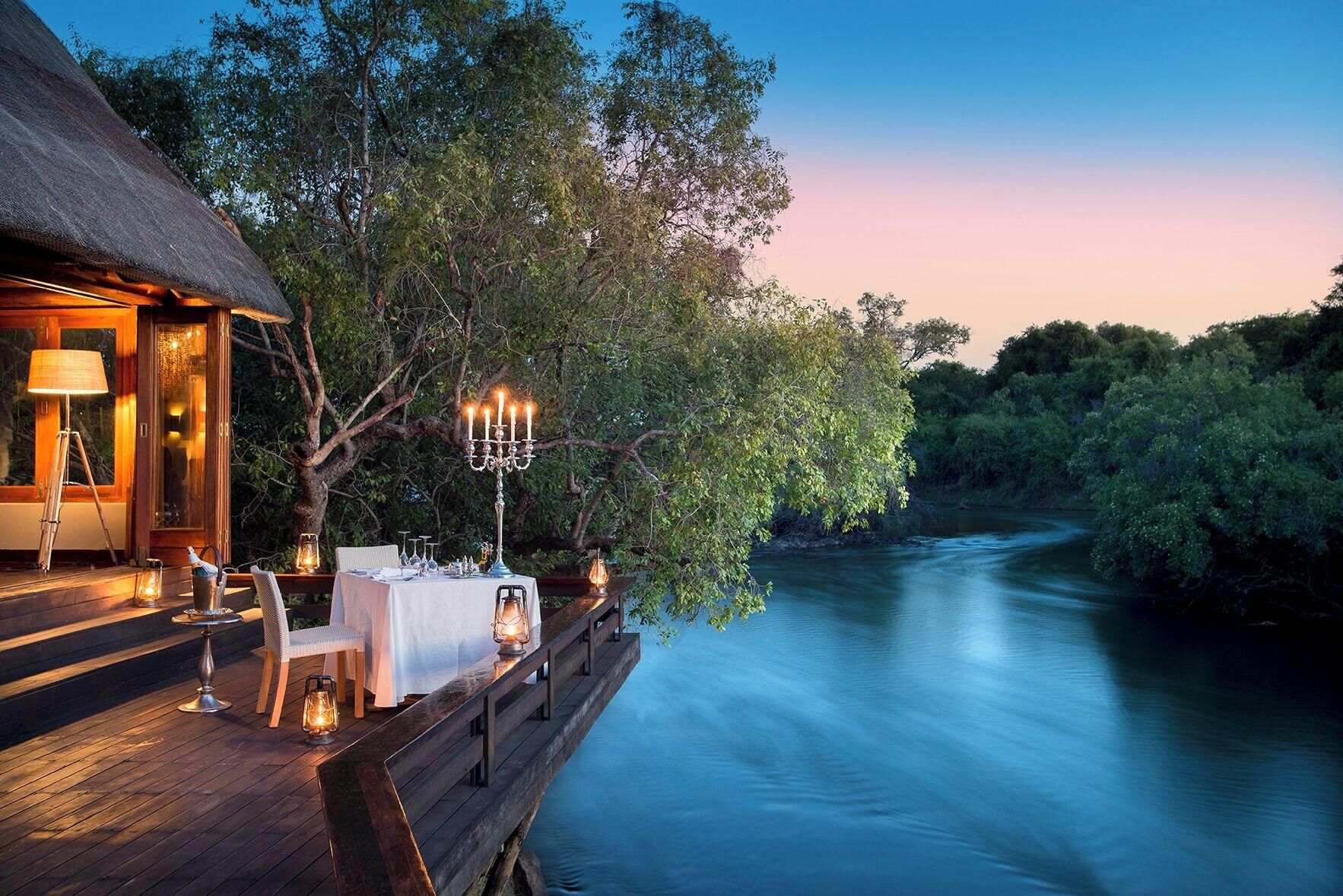 A dining area with a table set for two, overlooking a river, with a thatched-roof building to the left.