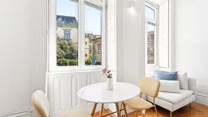 A room with a round white table, two beige chairs, a white sofa with blue cushions, and large windows with a view of buildings and greenery. - GuestReady - studio in Santa Catarina (Porto)