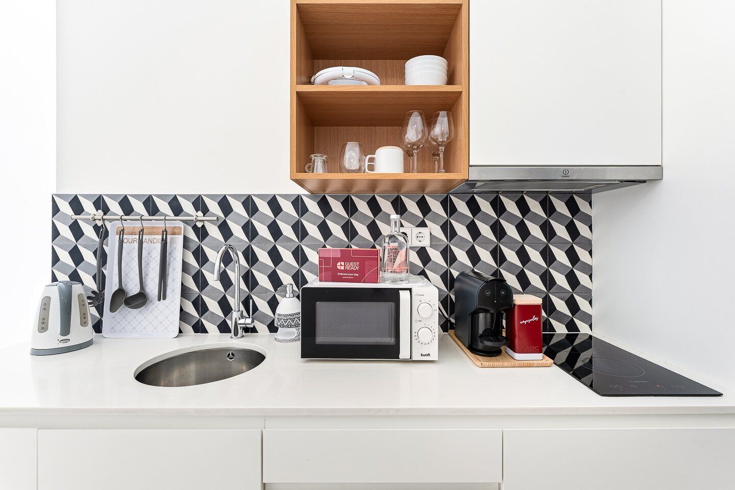 A modern kitchen with a microwave, toaster, kettle, and coffee maker. There is a wooden shelf above the counter holding plates and glasses.