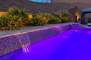 A swimming pool with a tiled edge and a waterfall feature, surrounded by landscaped greenery and a brick wall. - Three bedroom  house with beautiful pool (Lake Havasu City)