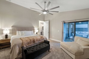 A bedroom with a bed, a black upholstered bench, a ceiling fan, a nightstand with a lamp, and a sliding glass door leading to a balcony. - Three bedroom  house with beautiful pool (Lake Havasu City)