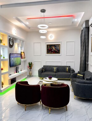 Ein modernes Wohnzimmer mit einem grauen Sofa, zwei schwarzen Sesseln, einem weißen Couchtisch und einem Flachbildfernseher an einer weißen Konsole.