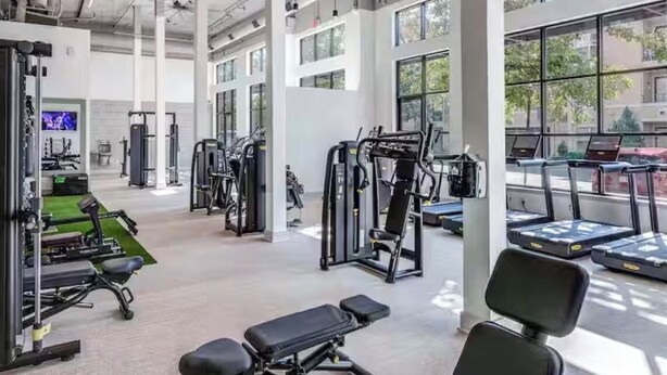 Ein Fitnessraum mit Trainingsgeräten, großen Fenstern und einem Deckenventilator.