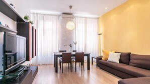 Un soggiorno moderno con una TV a schermo piatto, un tavolo da pranzo con sedie, un divano e un'ampia finestra con tende.