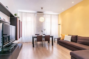 Ein modernes Wohnzimmer mit Flachbildfernseher, einem Esstisch mit Stühlen, einer Couch und einem großen Fenster mit Vorhängen.