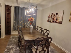 Uma sala de jantar com uma mesa de madeira, cadeiras, um candelabro, um vaso com flores, cortinas e um quadro na parede.