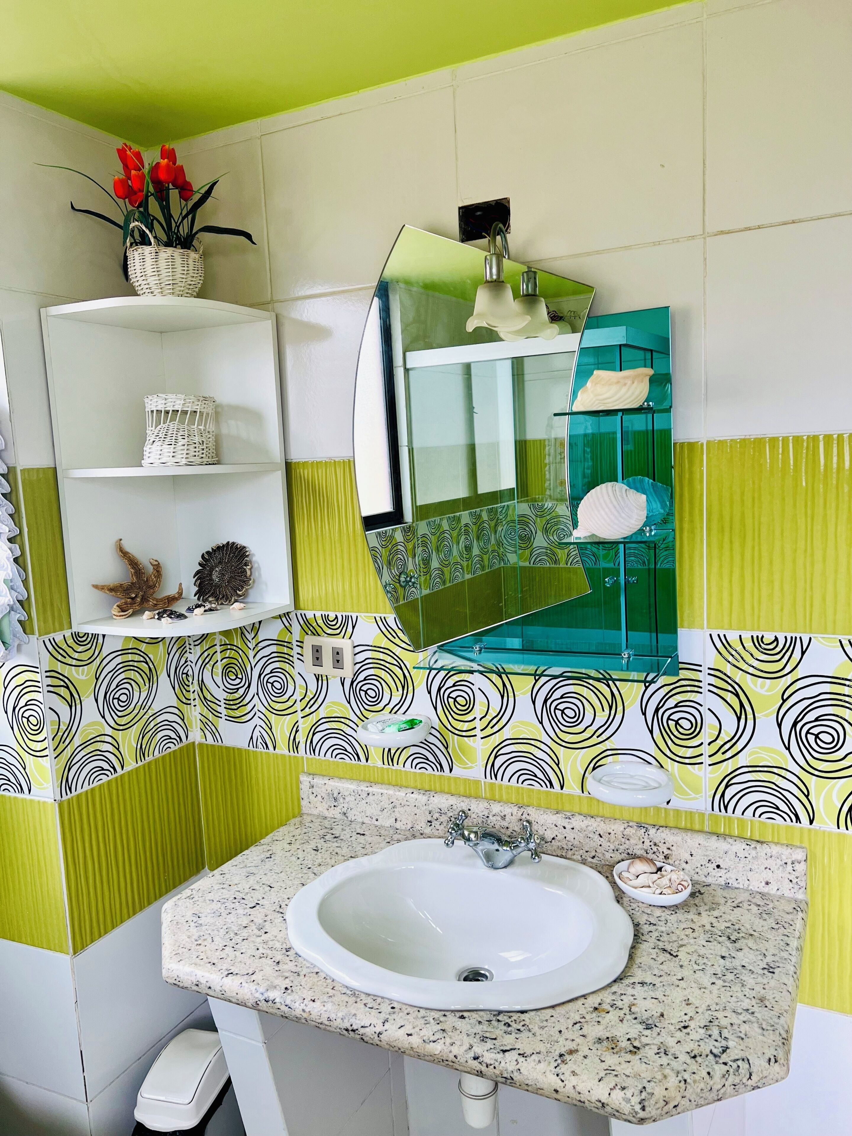 A bathroom with a white sink, green and white patterned tiles, a mirror, and a shelf with decorative items.