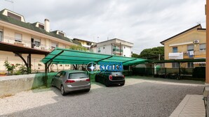 Ein Wohngebiet mit zwei geparkten Autos unter einer Baumkrone, ein Gebäude mit Balkonen und ein Fliesenboden.