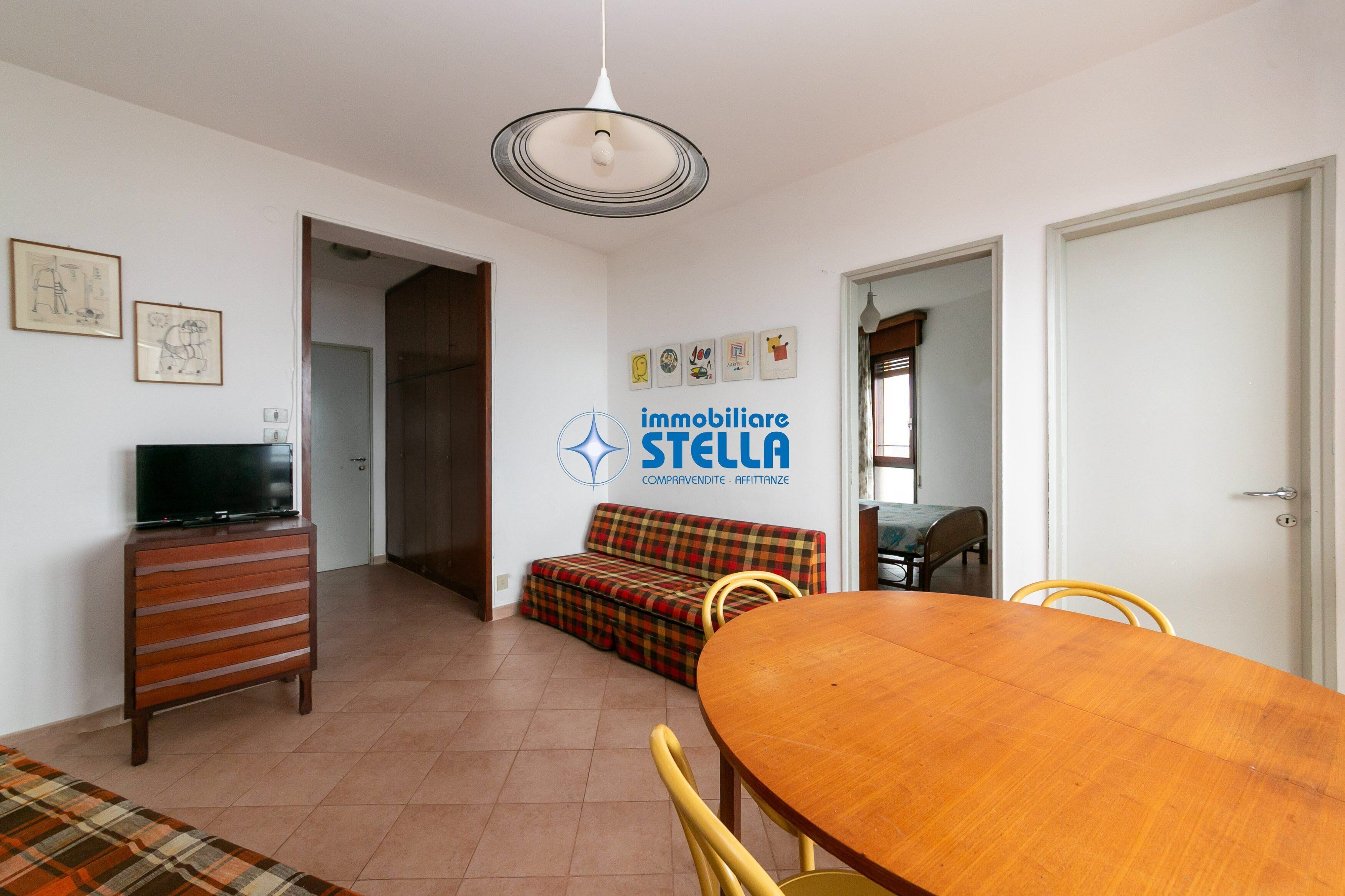 A dining area with a wooden table and chairs, a sofa, a television on a wooden cabinet, and a wall with a logo and pictures.