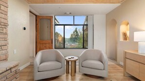 Una habitación moderna con dos sillones, una mesita redonda, un armario de madera y un ventanal con vistas a árboles y un edificio.