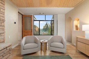A modern room with two armchairs, a small round table, a wooden cabinet, and a large window with a view of trees and a building. - Dry Creek Escape, Hot Tub, Views, Fireplaces (Sedona)