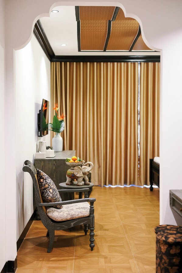 Une pièce avec un plancher en bois, une chaise en bois foncé ornée d’un coussin à motifs, une petite table sur laquelle se trouve un bol décoratif, et un grand rideau.