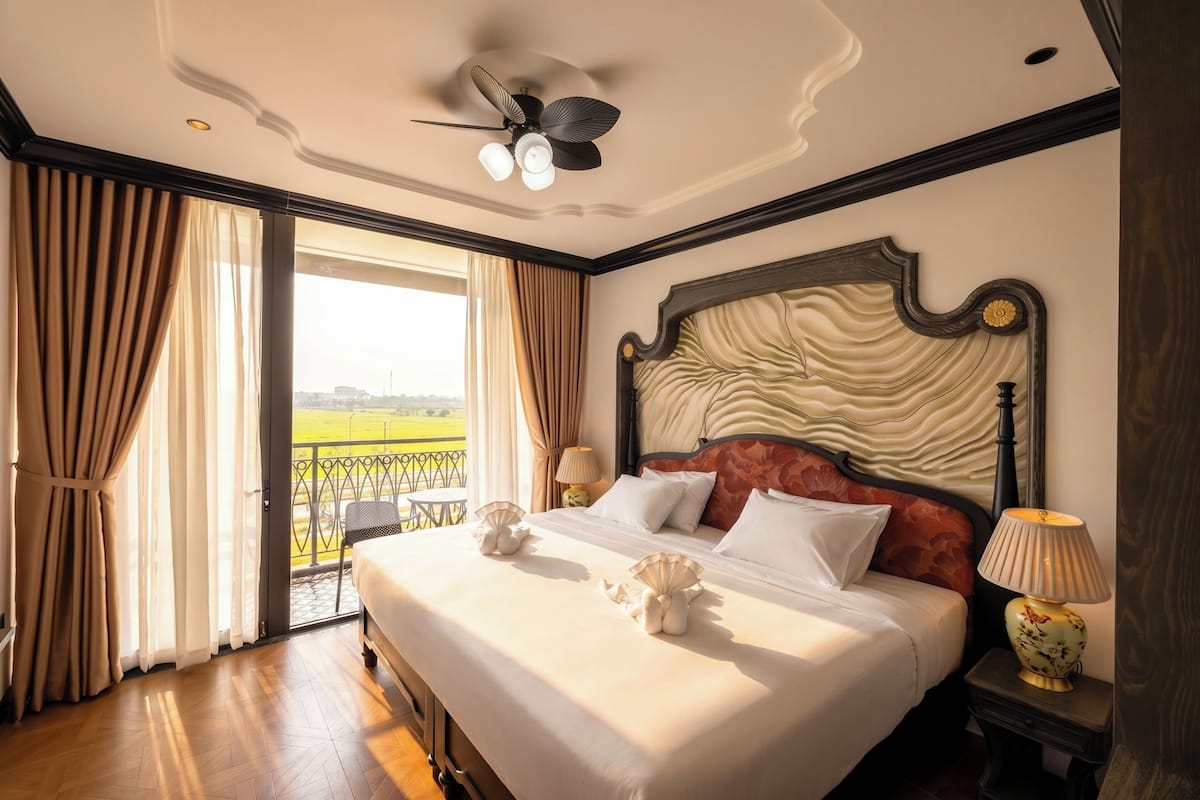 Une chambre d’hôtel avec un grand lit, un ventilateur de plafond, une table de chevet avec une lampe et une vue sur un paysage verdoyant, visible à travers une porte coulissante.