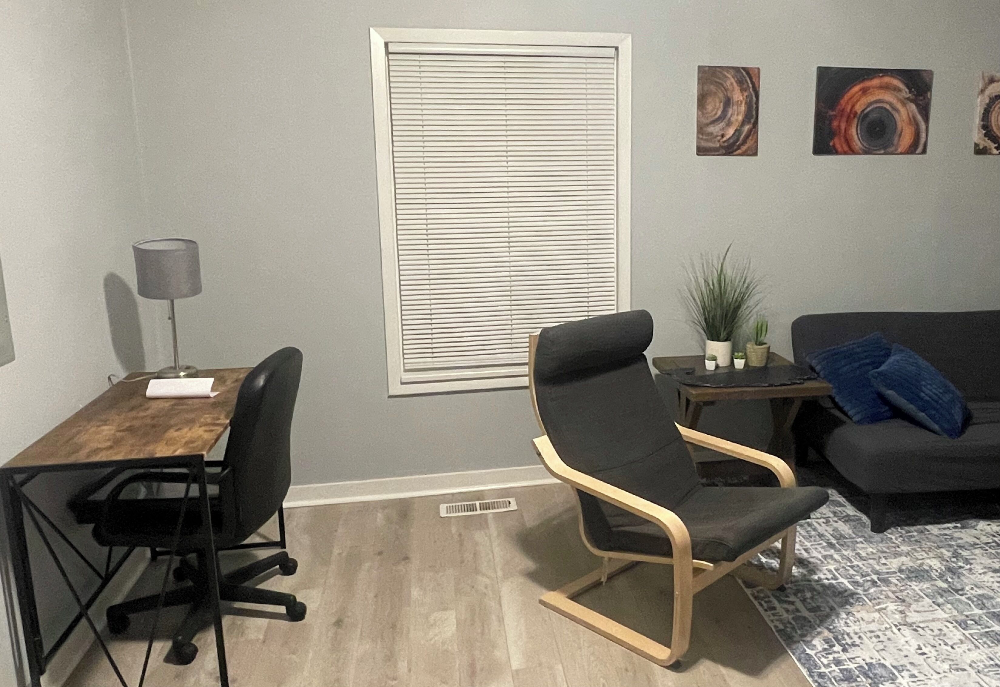A room with a wooden desk, black office chair, grey sofa, and a small side table with a plant.