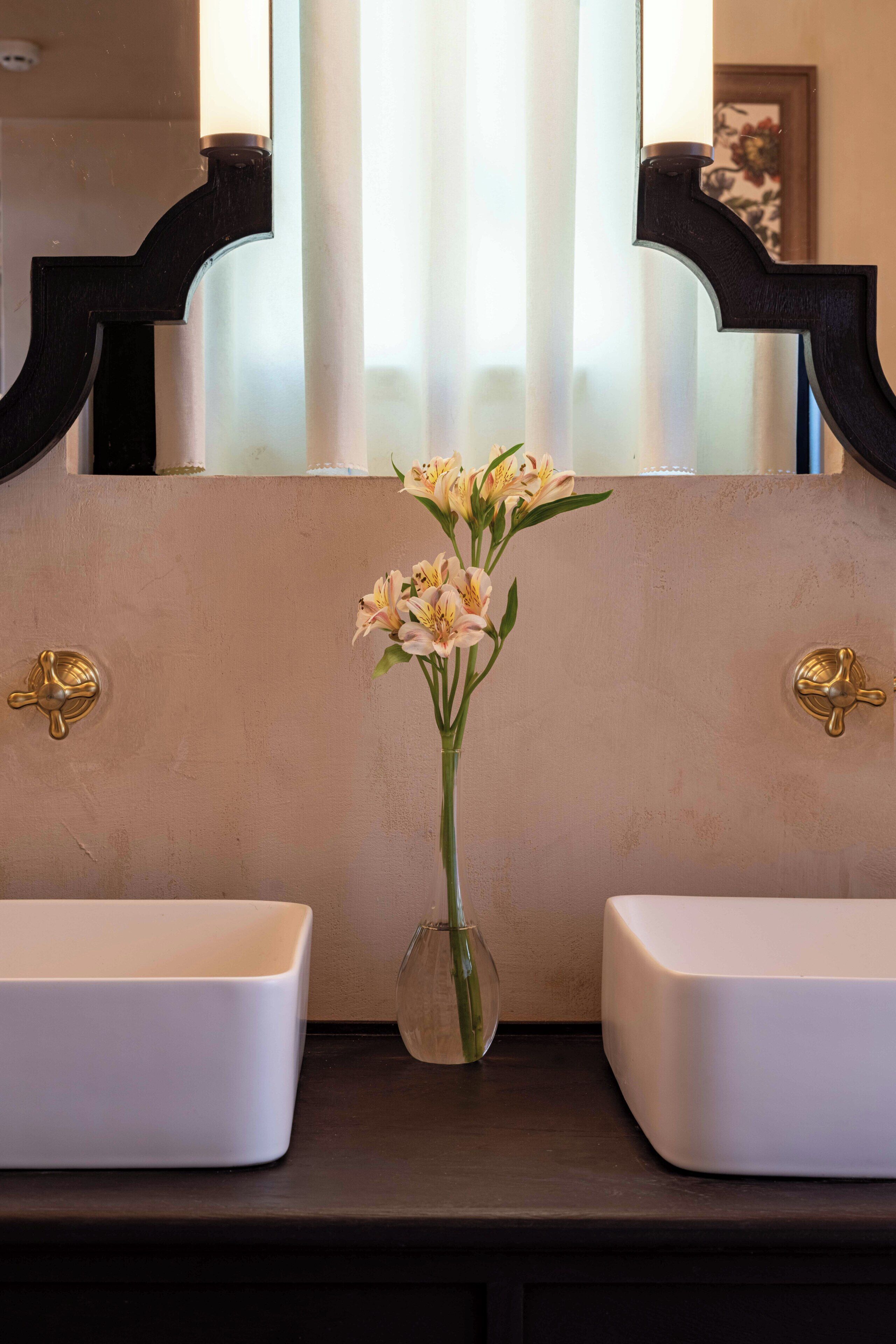Un baño con dos lavamanos blancos, un florero con flores y un espejo con marco decorativo.