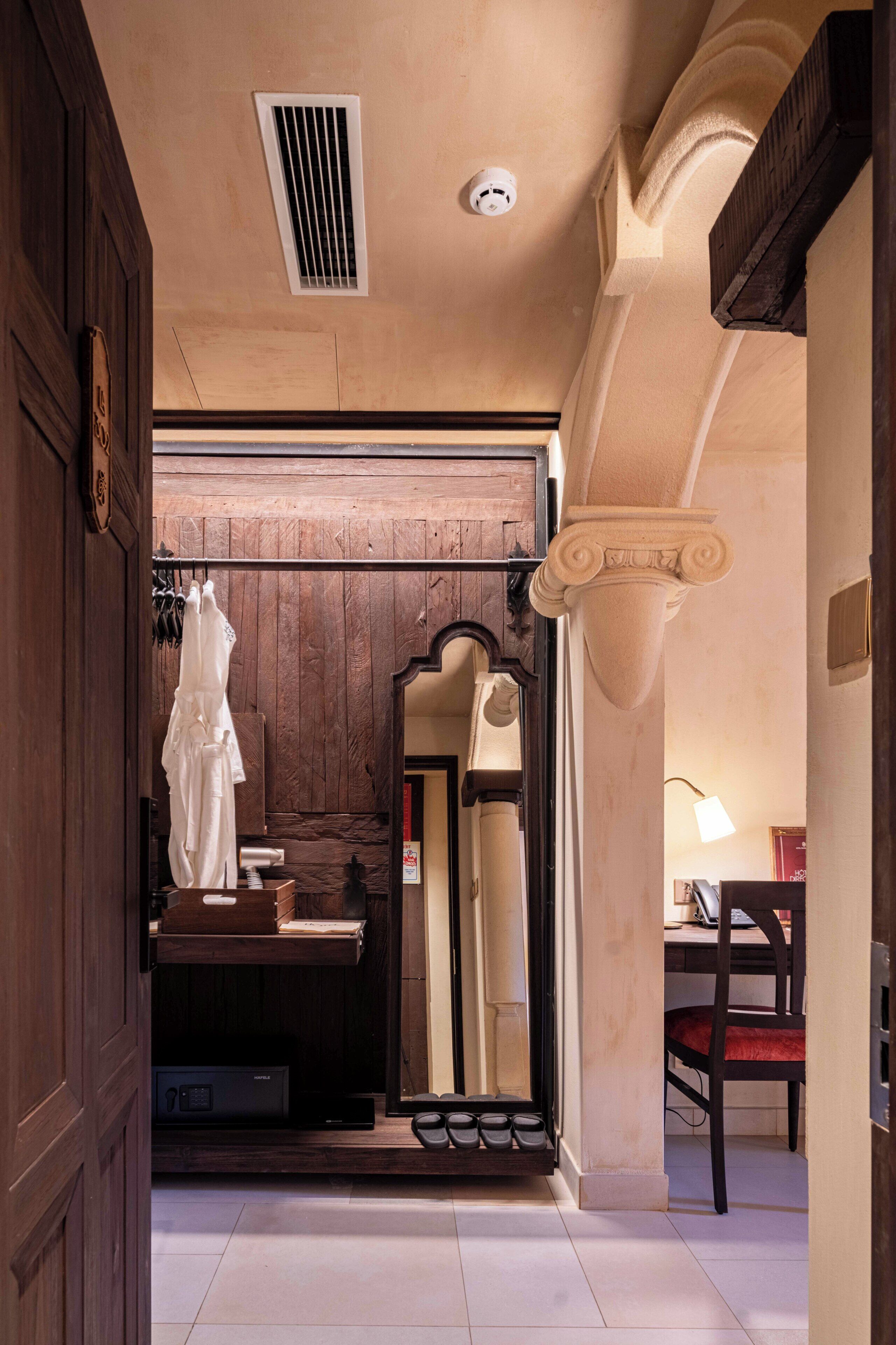 A room with a wooden door, a mirror, a white robe, a desk, and a chair.
