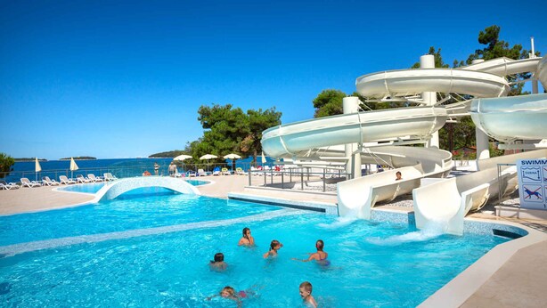 Una piscina con varias toboganes, sillas de descanso y sombrillas.