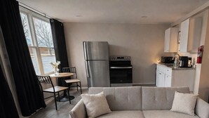 A modern living room with a sofa, a dining table with chairs, a kitchen area with a refrigerator and oven, and a large window with curtains. - Riverside apartment in Bay City perfect for your next getaway (Bay City)